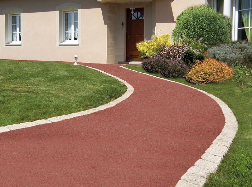 Une allée de jardin sinueuse en revêtement rouge et bordée de pavés clairs mène à l'entrée d'une maison.