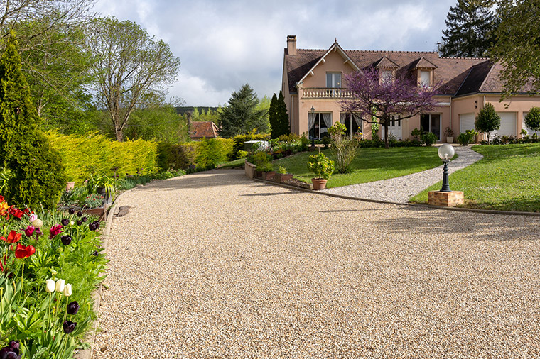 Une large allée de jardin en gravier beige mène à une maison de campagne entourée d'un jardin fleuri et de pelouses soignées.
