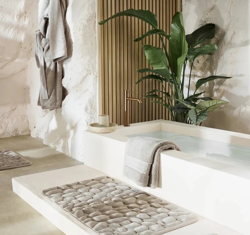 Une salle de bain avec une baignoire blanche, un tapis de bain à relief de galets et une grande plante verte décorative