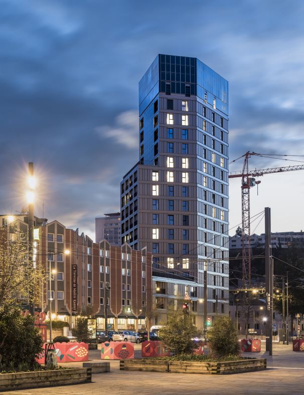 Immeuble moderne et haut en ville, éclairé le soir, avec des fenêtres allumées et des bâtiments autour.